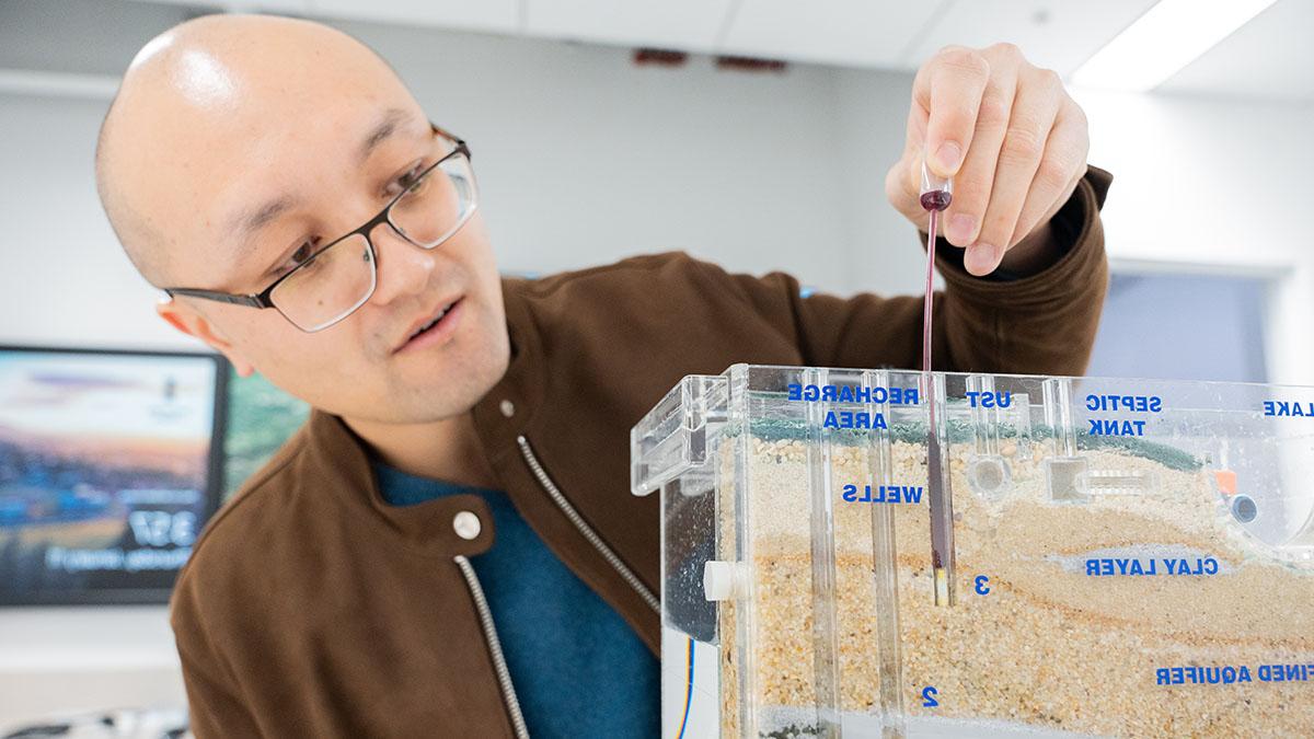 Meng Zhao, 一个戴着眼镜的秃顶男人，蓝色t恤外穿一件棕色夹克, 将一根小棒插入一个塑料罐中，罐中有不同层的标记土壤. 